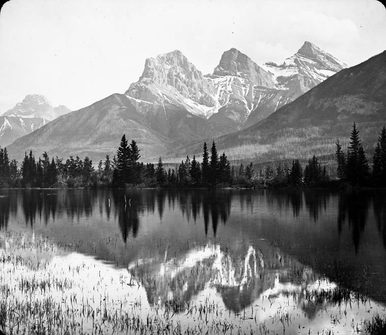 Photo en noir et blanc de trois majestueux sommets enneigés qui se reflètent dans le lac au premier plan.