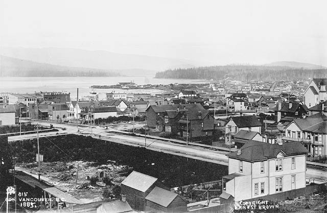 Photo en noir et blanc d’une nouvelle ville côtière avec l’océan et les montagnes au fond. Les premiers bâtiments à un ou deux étages, une grande parcelle inoccupée et des routes se trouvent au premier plan.