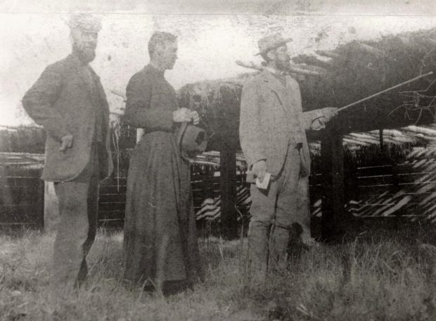 Photo en noir et blanc de trois hommes à l’extérieur. Deux d’entre eux portent un costume et l’homme de droite pointe une canne. L’homme au centre porte une soutane et tient un chapeau.