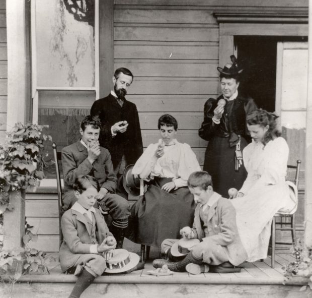 Photo en noir et blanc montrant une famille assise sur une véranda : un homme, deux femmes, une fille et trois garçons qui regardent tous la pomme qu’ils tiennent à la main.