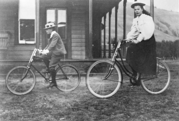 Photo en noir et blanc d’une fille et d’un jeune garçon faisant du vélo près d’une maison.