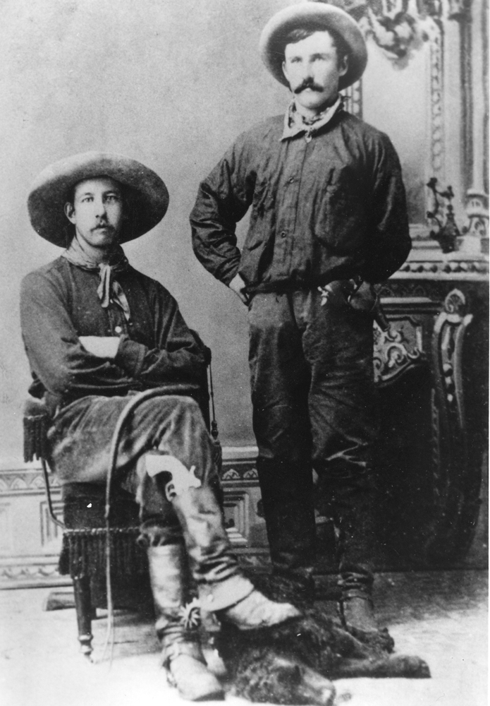 Photo en noir et blanc de deux hommes en tenues et chapeaux de cowboy. L’un d’eux est assis, l’autre debout.
