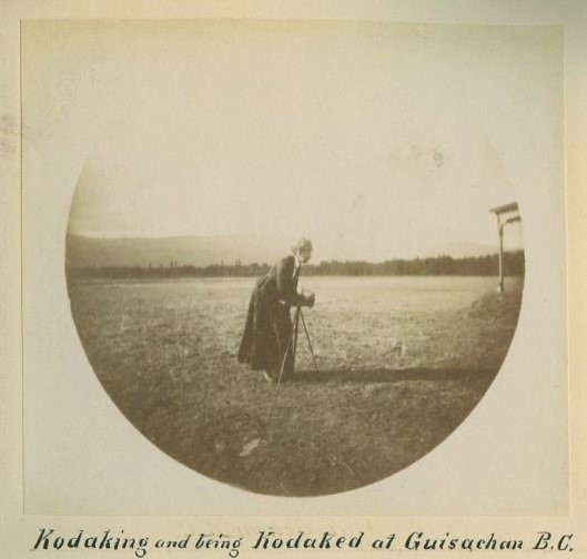 Photo ronde en noir et blanc montrant une femme dans un champ avec un vieil appareil photo sur un trépied.