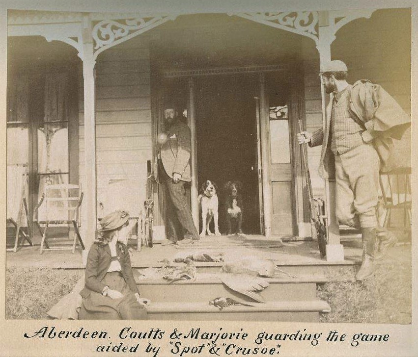 Photo en noir et blanc de deux hommes, une fille, deux chiens et des oiseaux morts sur une galerie. L’homme de droite tient un fusil.