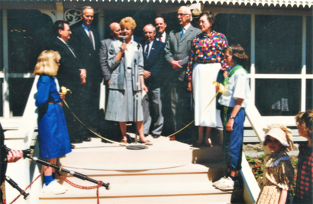 Photo en couleur représentant un groupe de personnes debout sur une véranda. Au milieu, une femme parle dans un micro.