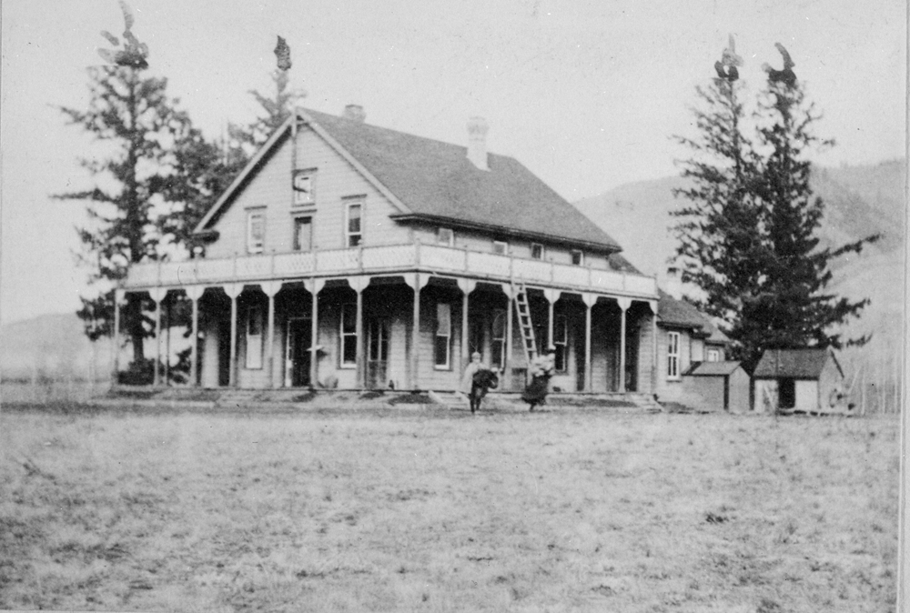 Photo en noir et blanc de la façade et du côté d’une maison de deux étages entourée d’une véranda et agrémentée d’un balcon. On voit deux enfants debout devant la maison.