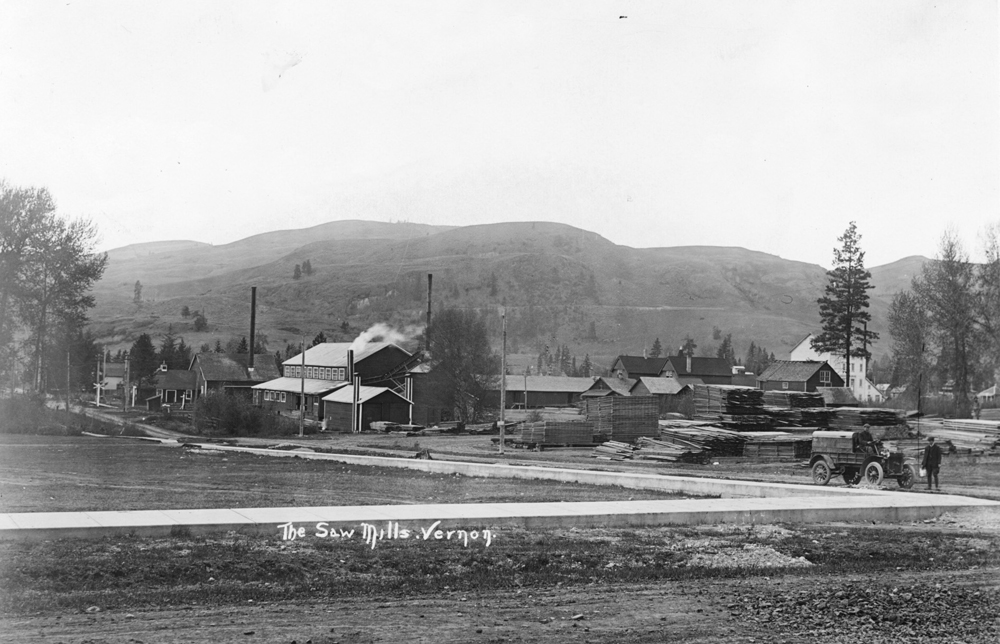 Photo en noir et blanc montrant trois petits édifices industriels en bois près d’une route avec des collines en arrière-plan.