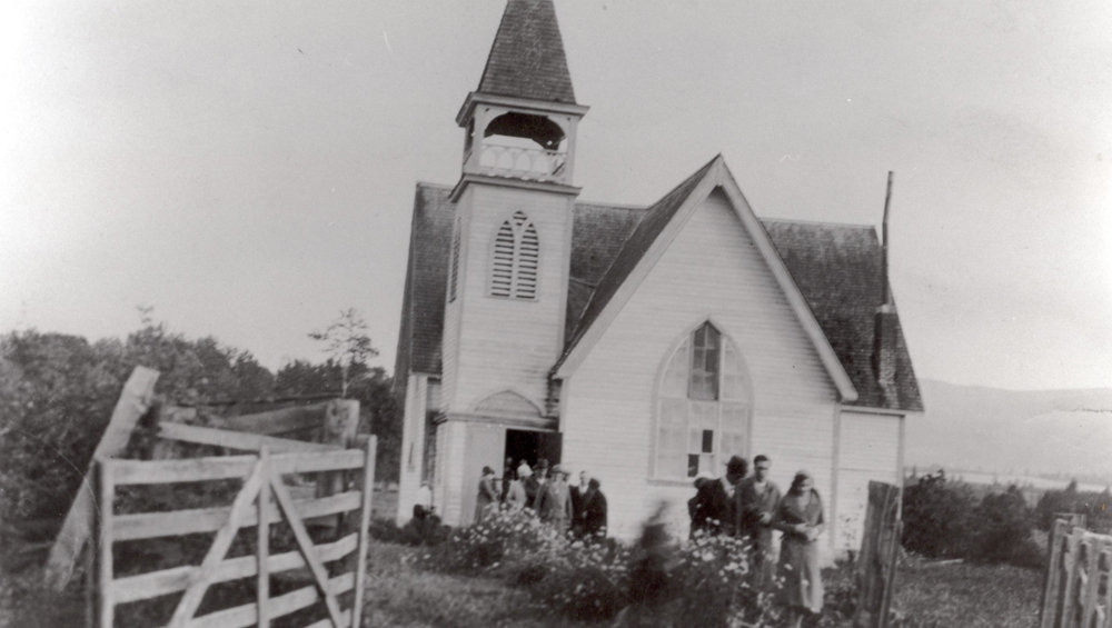 Photo en noir et blanc représentant un groupe d’hommes et de femmes sortant d’une petite église.