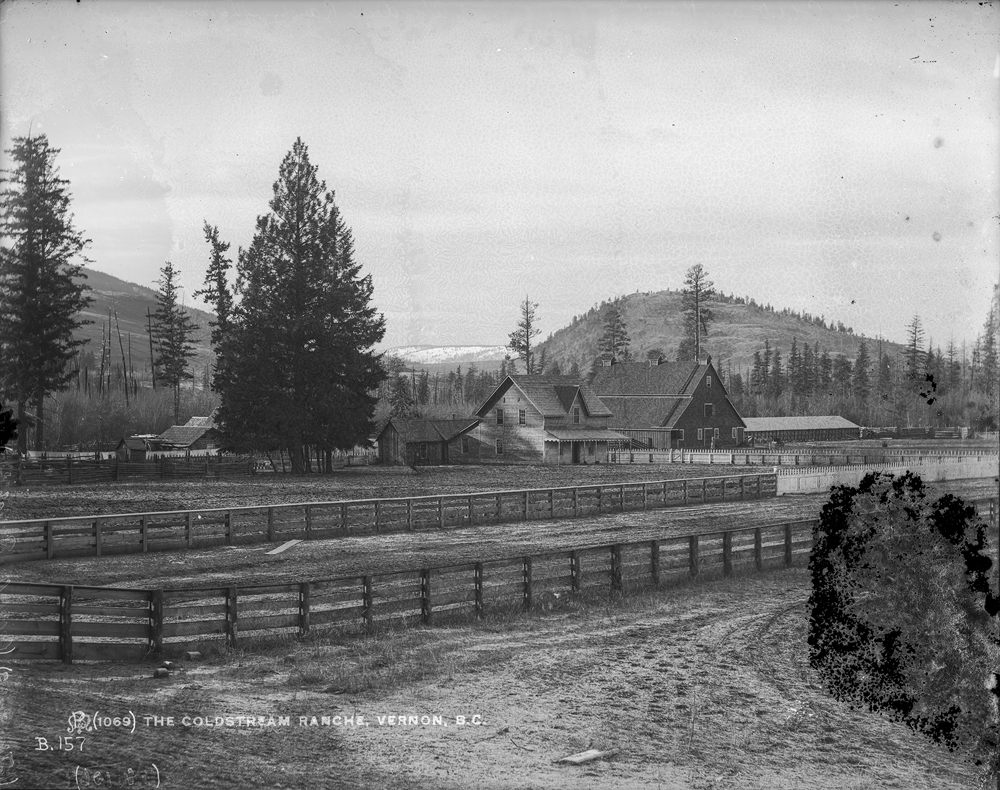 Photo en noir et blanc d’une clôture de ferme devant des granges et des arbres, avec des collines au loin.