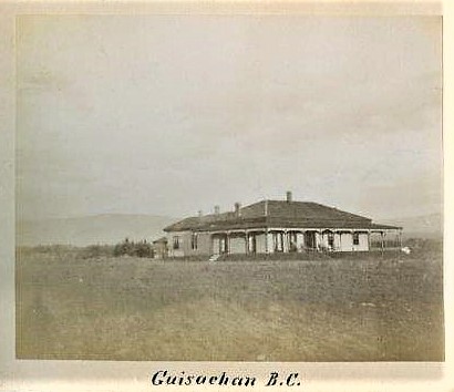 Photo en noir et blanc montrant la façade et le côté d’une maison à étage entourée d’une véranda avec un champ devant.