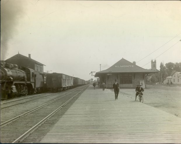 Photo de la gare d’Almonte avec un train sur la voie ferrée au loin. Des hommes sont debout sur le quai et un homme se promène à vélo à côté du quai, années 1930