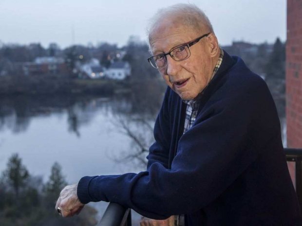 Photographie en couleur de Douglas Snair dans sa maison, 2017; on le voit debout sur son balcon avec un plan d’eau à l’arrière. 