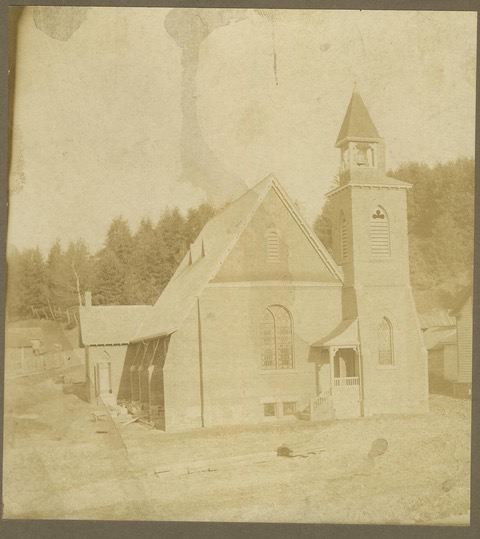 Photo en noir et blanc, et très jaunie, de l'actuel St. James the Apostle. La photo a été prise en 1902 lors de l'ouverture. La photo montre l'église, le portique d'entrée et le clocher. En arrière-plan, on voit la colline boisée des églises précédentes.