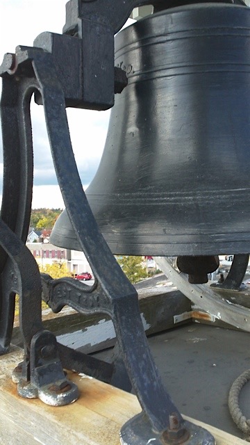 Photographie couleur de la cloche sur le clocher de l'actuelle église St. James. La cloche est peinte d'une couleur grise. À l'arrière-plan, on voit une vue de la ville de Fenelon Falls, avec le lac Cameron au fond de l'image.