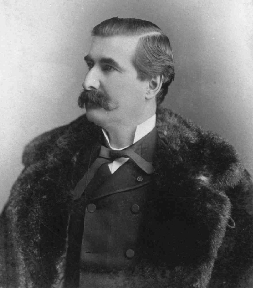 Photographie noir et blanc d’un homme dans la quarantaine. Ses cheveux sont foncés et il arbore une moustache fournie. Sous un manteau de fourrure, l’homme porte des vêtements élégants.