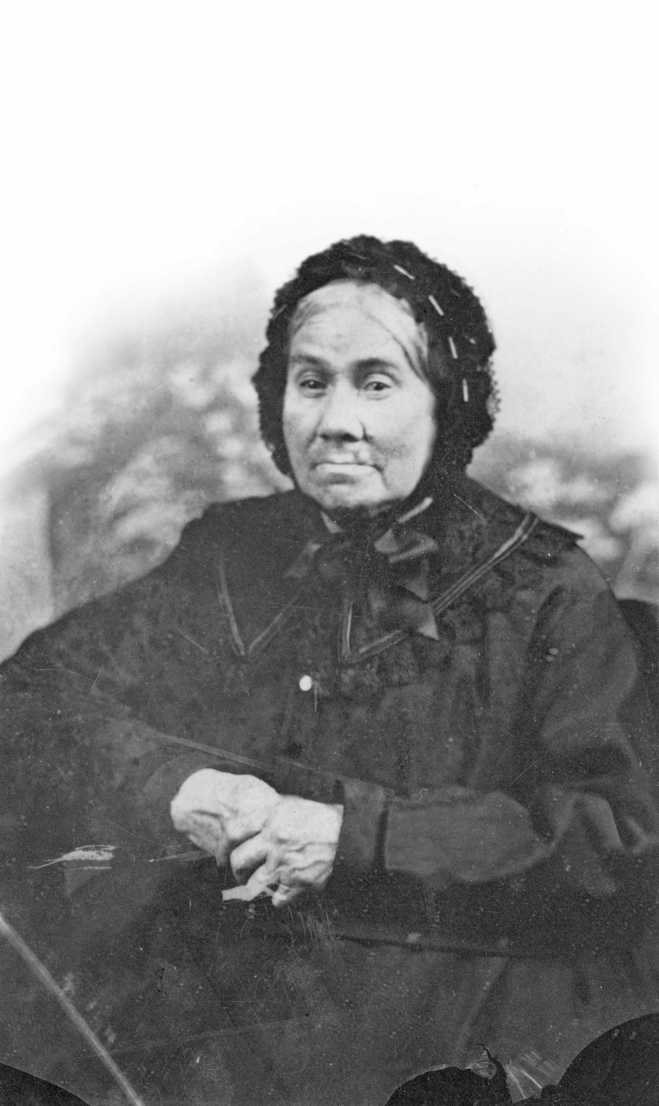 Photographie sépia d’une femme âgée. Celle-ci porte une longue robe noire ornée d’un large collet de dentelle. Ses cheveux blancs attachés à l’arrière de la tête en chignon sont couverts par un bonnet (un béguin) de dentelle noire attachée sous le menton par un large ruban. Ses mains, croisées devant elle, montrent des doigts tordus et usés par la vieillesse.