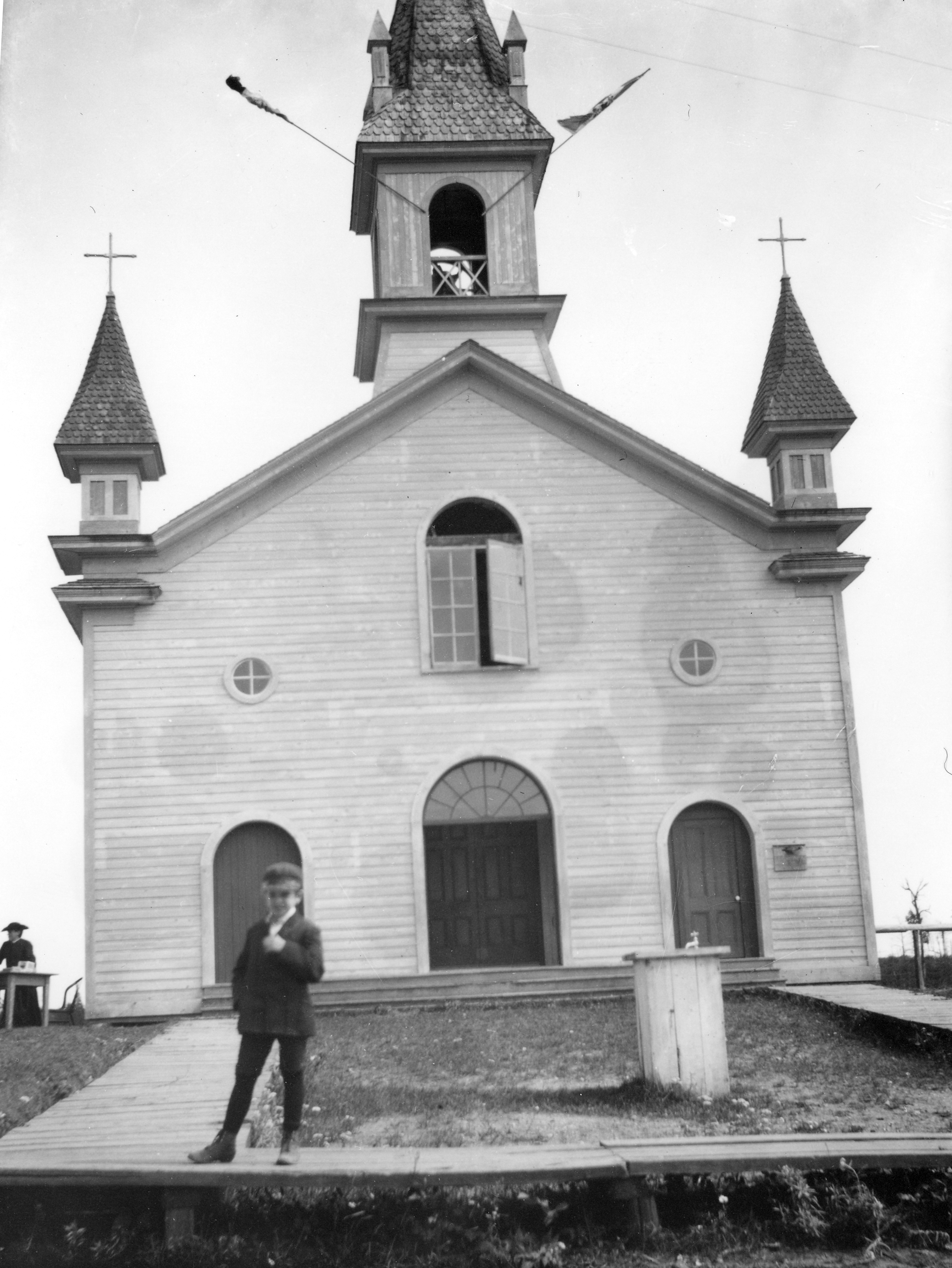 Photographie noir et blanc de la façade d’une petite église. Le bâtiment, dont le revêtement est en planches de bois, a trois portes, trois fenêtres au-dessus des portes et un clocher. Un jeune garçon se tient debout devant l’église sur le trottoir de bois.