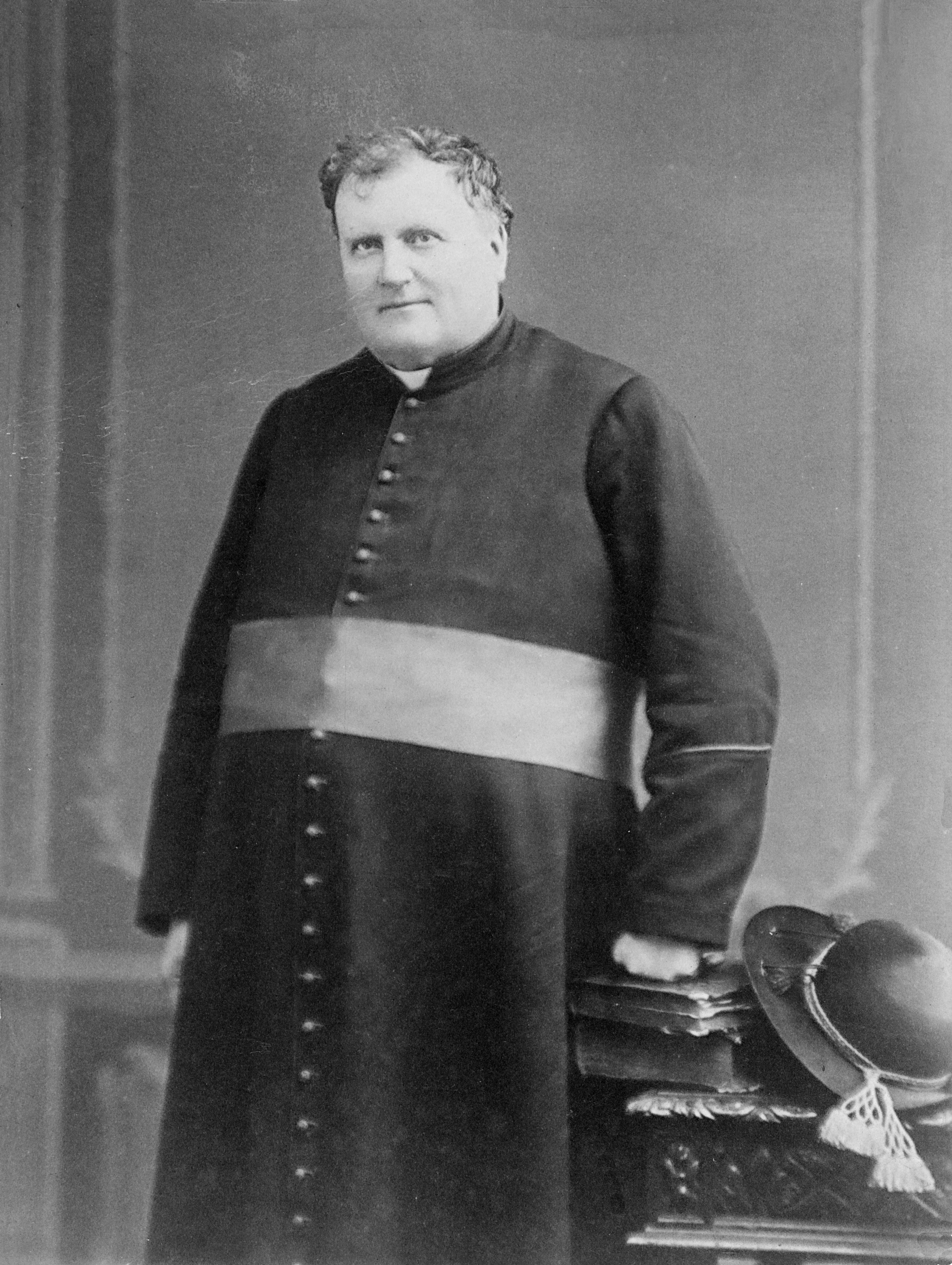 Photographie noir et blanc d’un homme dans la fin cinquantaine. L’homme est debout et porte une soutane. Une ceinture entoure sa taille, signe de son statut de monseigneur. Deux livres et un chapeau de feutre sont posés sur une table basse ornementée près de l’homme. L’homme pose un poing sur les livres.