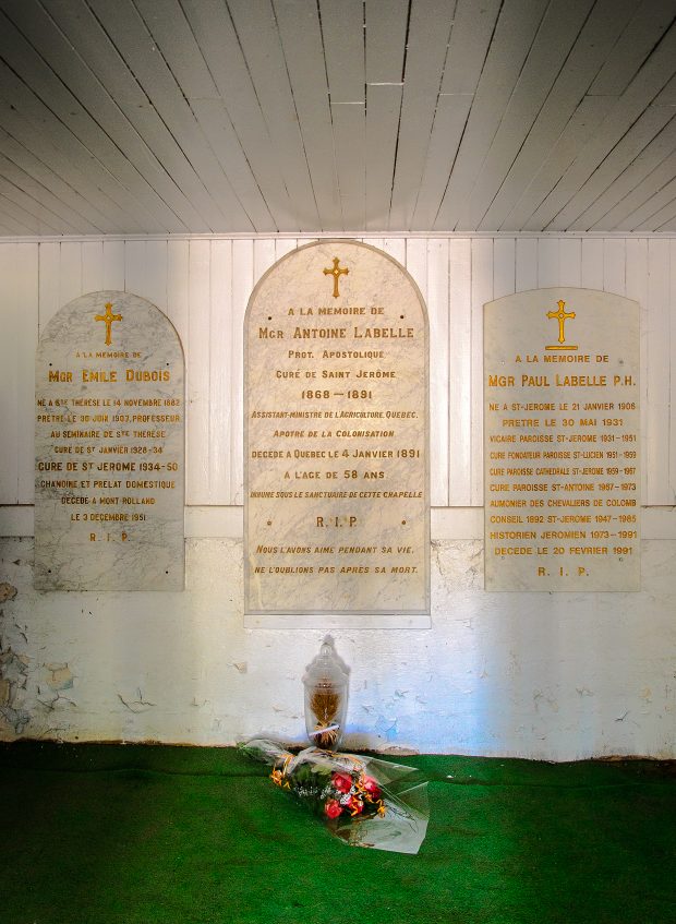 Photographie couleur d’une sépulture dans les sous-bassement d’une chapelle. Trois plaques de marbre blanc avec des écritures dorées sont accrochées au mur. Devant la plaque centrale, un bouquet de fleurs et une urne en verre sont déposés sur le sol recouvert de tapis gazon. 