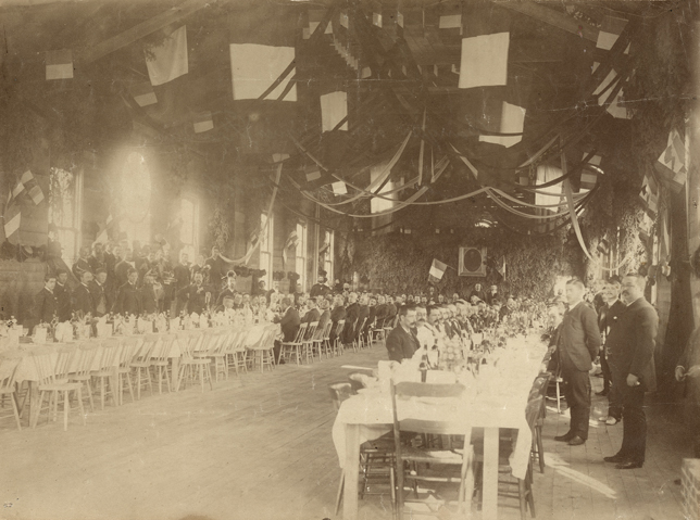 Photographie sépia d’une salle de réception plus longue que large et au plafond élevé. La salle est décorée de banderoles, de drapeaux français et de sapinage. On retrouve deux longues tables parallèles dressées pour un repas. Plusieurs hommes sont attablés, les membres d’une fanfare se tiennent debout près du mur gauche. 