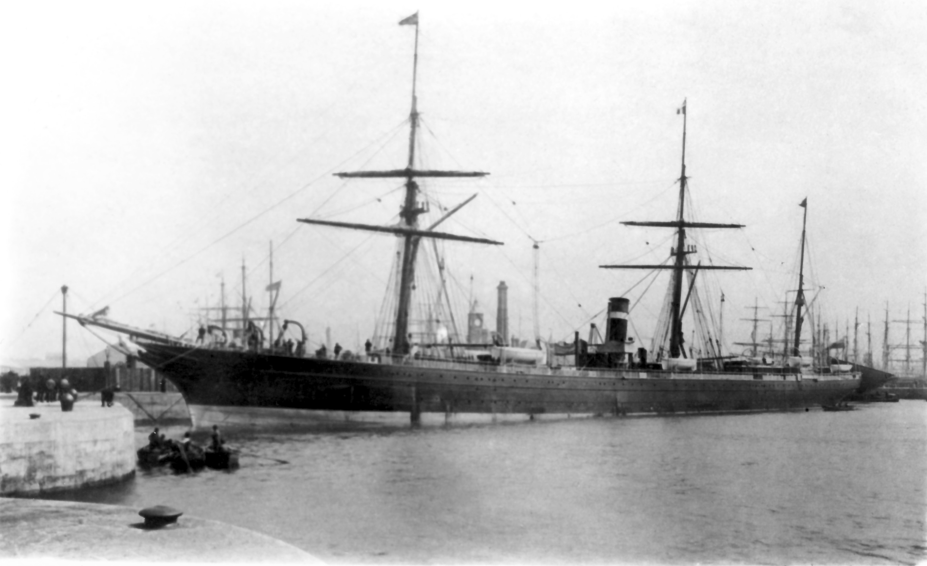 Photographie noir et blanc d’un bateau à voile et à vapeur accosté à un quai en béton dans un port. Le bateau, très grand, comprend trois mâts et une cheminée. Quelques chaloupes sont dans l’eau non loin du bateau. En arrière-plan, plusieurs mâts d’autres bateaux.