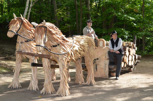 Photographie couleur montrant une sculpture réalisée par l’assemblage de bois de grève. L’œuvre de grandeur nature se trouve dans un boisé. Elle représente un traîneau rempli de billots de bois tiré par deux chevaux. Un homme sculpté dans le bois est assis à l’avant du traîneau. Deux hommes vêtus à la mode de l’époque prennent la pose sur le traîneau.