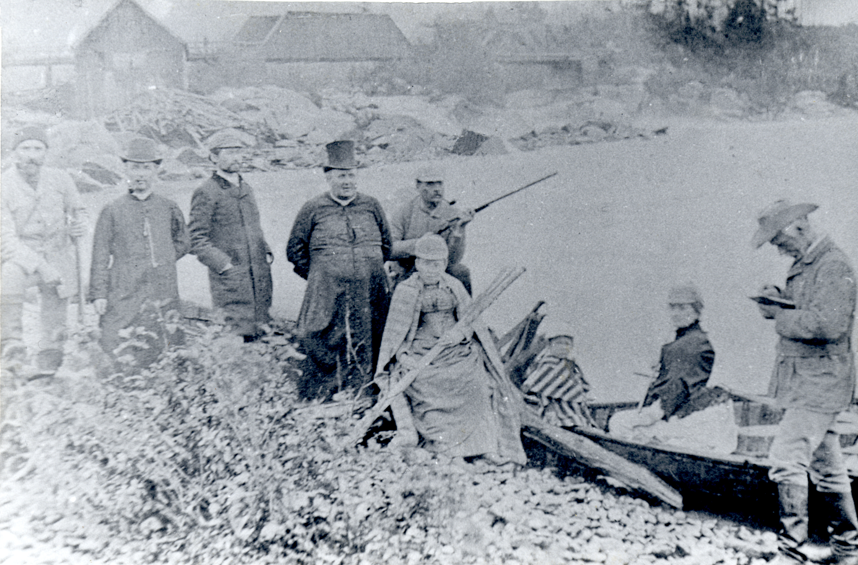 Photographie noir et blanc représentant un groupe de personnes sur le bord d’une rivière à côté d’un canot. On retrouve deux prêtres, deux femmes, un enfant, deux hommes tenant des carabines, un homme prenant des notes et un autre homme. En arrière-plan, on aperçoit un pont de bois et des bâtiments en bois.