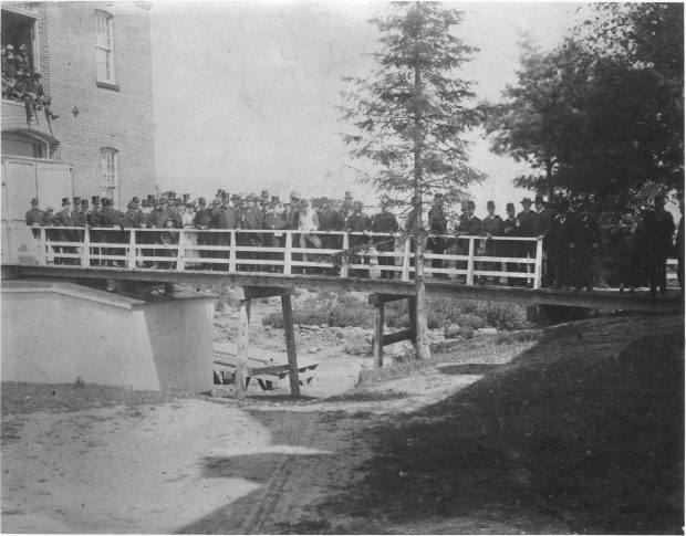 Photographie noir et blanc d’un groupe d’individus, majoritairement des hommes, debout sur une passerelle en été. La passerelle donne accès à un grand bâtiment de brique. On voit aussi des gens juchés plus haut dans une ouverture de l’usine. Plusieurs individus portent un chapeau haut de forme pour l’occasion.