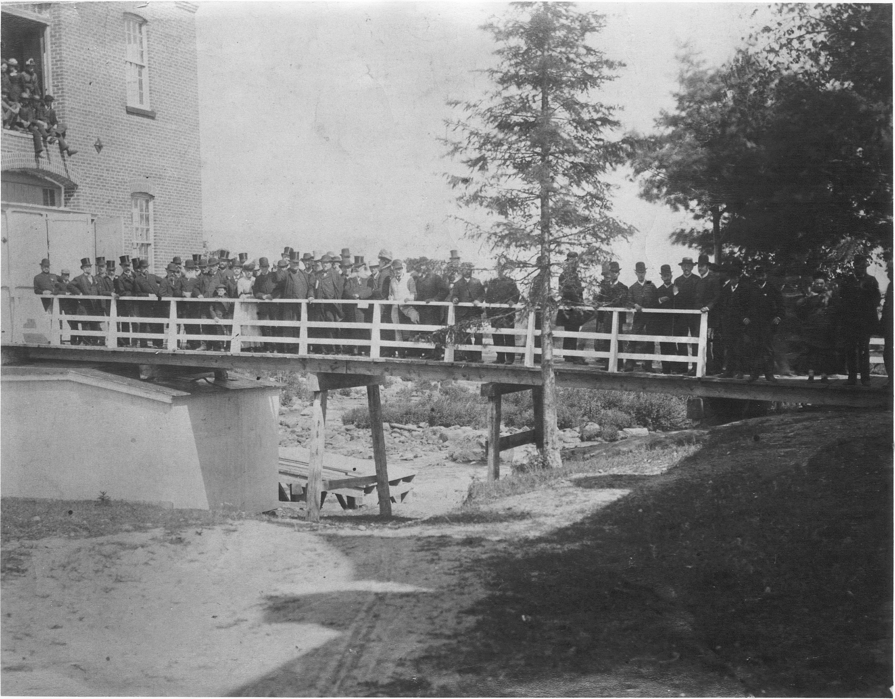 Photographie noir et blanc d’un groupe d’individus, majoritairement des hommes, debout sur une passerelle en été. La passerelle donne accès à un grand bâtiment de brique. On voit aussi des gens juchés plus haut dans une ouverture de l’usine. Plusieurs individus portent un chapeau haut de forme pour l’occasion.