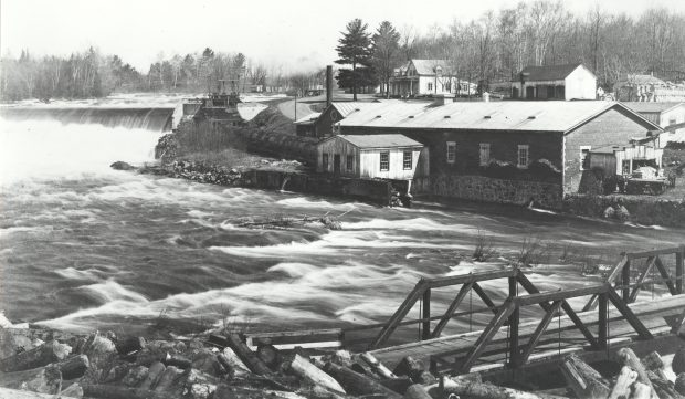 Photographie en noir et blanc d’un paysage au printemps. Au premier plan à droite, un petit pont de bois enjambant une rivière houleuse et un amoncellement billots de bois. En amont, un barrage d’où l’eau s’écoule avec force. Au centre, un bâtiment industriel de brique. En arrière-plan à gauche, une maison et quelques bâtiments de ferme.