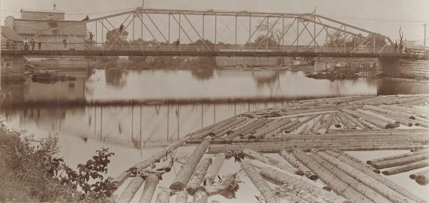 Photographie sépia d’un paysage en été dont le sujet principal est un pont de fer et une rivière. Quelques individus se tiennent sur le pont ou sont montés dans la structure. En premier plan, on aperçoit des troncs d’arbres qui flottent dans l’eau. Quelques bâtiments de bois sont en arrière-plan.