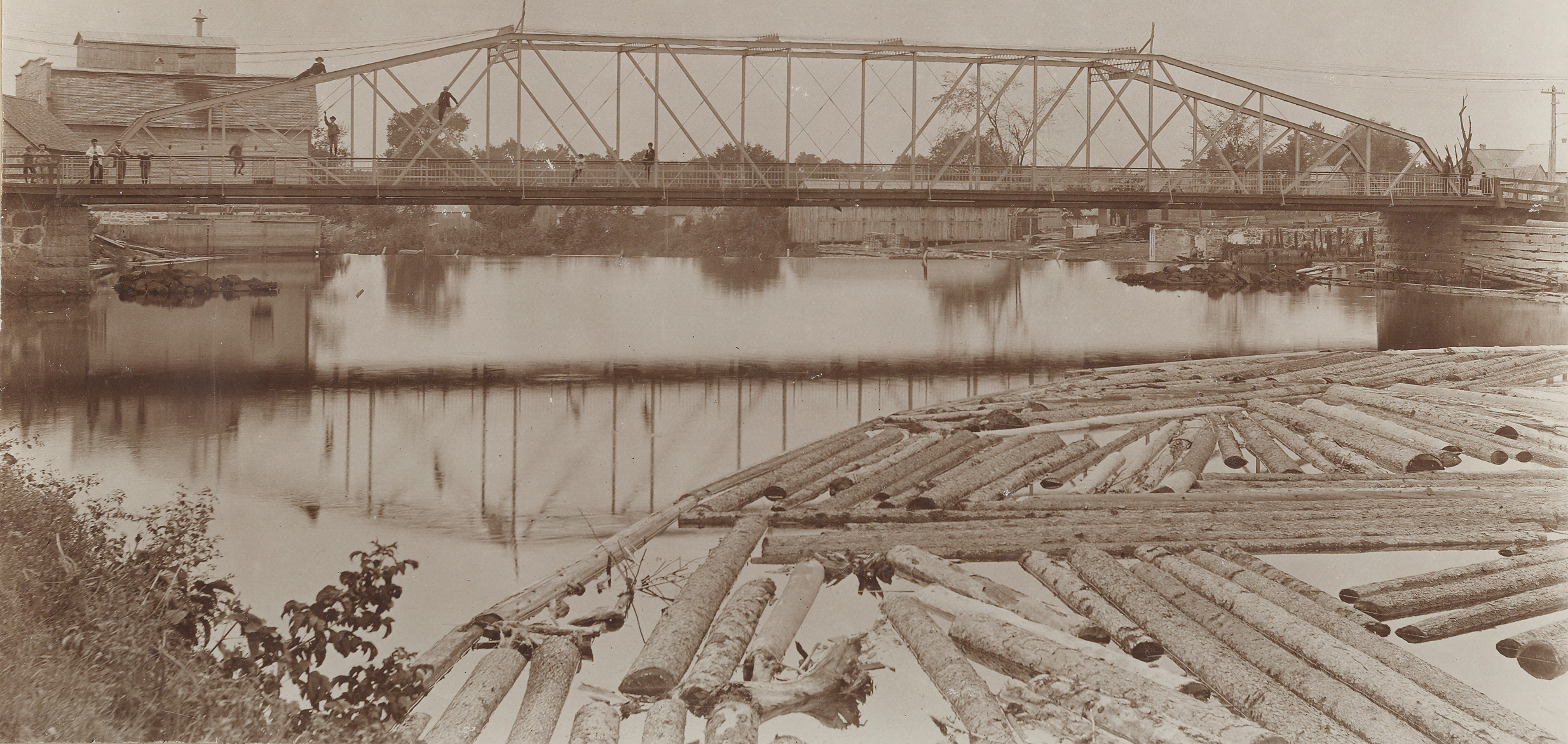 Photographie sépia d’un paysage en été dont le sujet principal est un pont de fer et une rivière. Quelques individus se tiennent sur le pont ou sont montés dans la structure. En premier plan, on aperçoit des troncs d’arbres qui flottent dans l’eau. Quelques bâtiments de bois sont en arrière-plan.