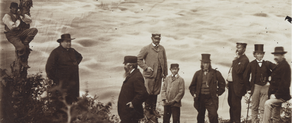 Photographie noir et blanc d’un groupe de neuf personnes sur la rive d’un cours d’eau tumultueux. À gauche, un homme est suspendu au tronc d’un arbre. Le curé Labelle, homme corpulent, porte une soutane et un chapeau de feutre à large bord. Au centre et à droite, six hommes et un adolescent en habit trois-pièces portant des chapeaux hauts de forme, casquettes ou chapeaux de feutre. Deux d’entre eux fument la pipe.