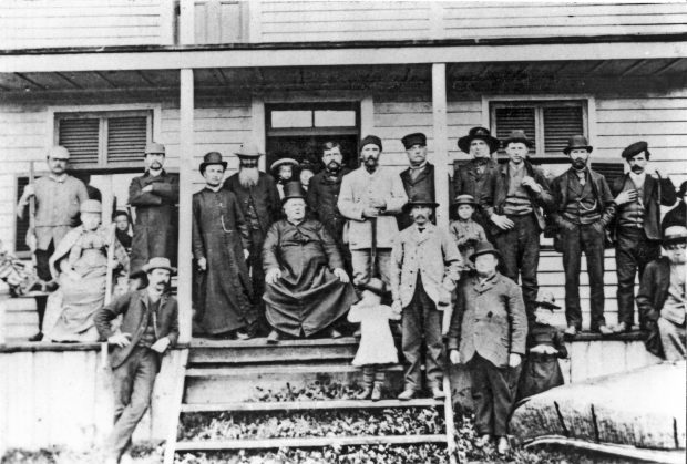 Photographie d’un groupe d’une vingtaine d’individus sur la galerie d’une maison en bois. Au centre du groupe, le curé Labelle, un homme corpulent en soutane est assis sur une chaise. À droite, sur le sol devant la galerie, un grand canot d’écorce.