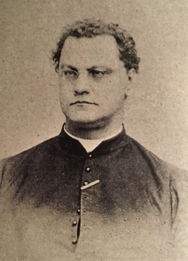 Photographie sépia d’un homme dans la trentaine. Il a les cheveux foncés, courts et bouclés. Il porte de petites lunettes ovales. Il est vêtu d’une soutane et un col romain blanc entoure son cou. 