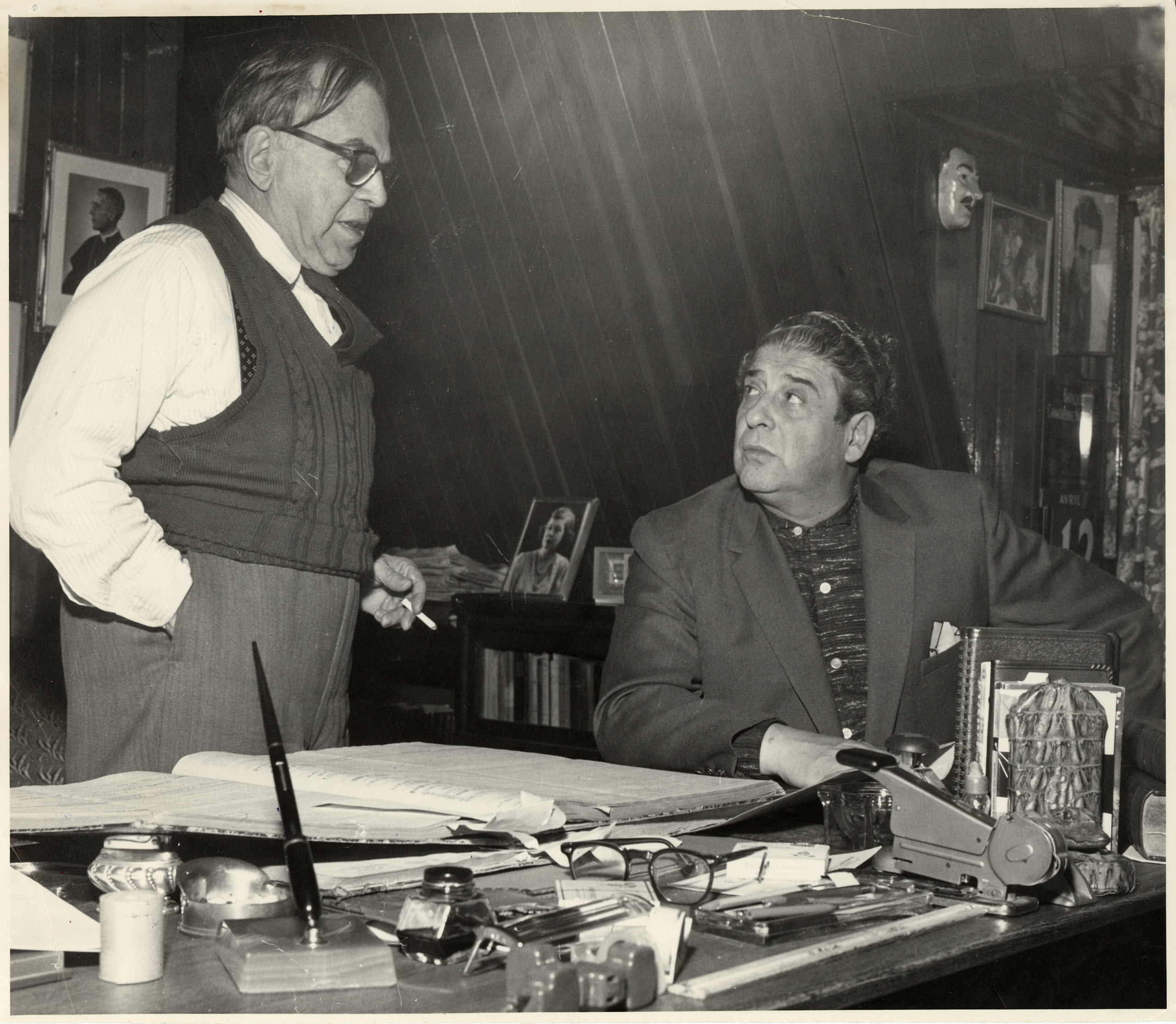 Un homme est debout près d’un bureau et discute avec un autre homme assis près de lui. Le bureau est chargé. On y trouve un grand livre ouvert, une brocheuse, des lunettes, un encrier, une plume et une multitude d’autres petits objets.