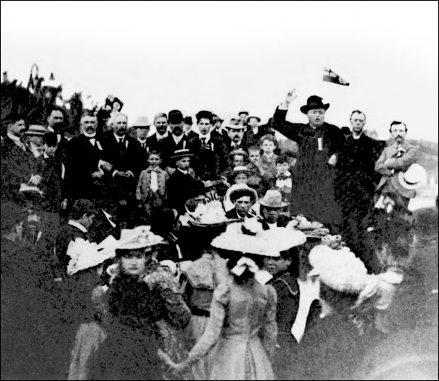 Photographie noir et blanc d’une foule à l’extérieur entourant un homme imposant, le curé Antoine Labelle. On aperçoit plusieurs hommes, quelques garçons et quelques femmes tous bien vêtus. Il prononce un discours ; sa bouche est ouverte et son bras droit est levé près de sa tête. Son bras gauche est posé sur sa hanche. Il porte une soutane noire et un chapeau noir. 