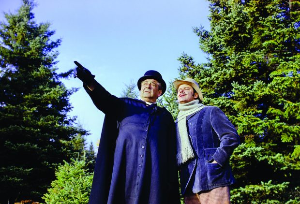 Photographie couleur de deux comédiens au milieu de la forêt. L’homme en soutane, qui représente le curé Labelle, pointe son doigt devant lui. L’homme qui l’accompagne est près de lui et regarde au loin. Il porte un manteau, un foulard et un chapeau. 