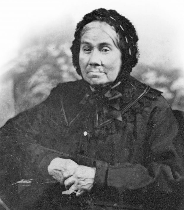 Photographie sépia d’une femme âgée. Celle-ci porte une longue robe noire ornée d’un large collet de dentelle. Ses cheveux blancs attachés à l’arrière de la tête en chignon sont couverts par un bonnet (un béguin) de dentelle noire attachée sous le menton par un large ruban. Ses mains, croisées devant elle, montrent des doigts tordus et usés par la vieillesse. 