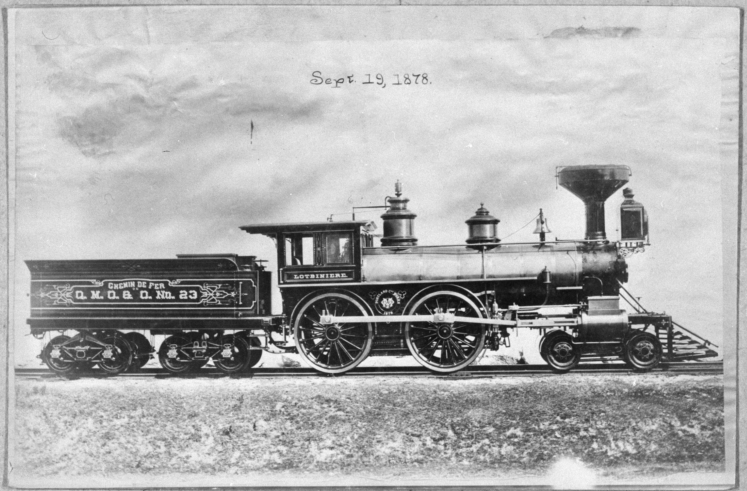 Photographie noir et blanc d’une locomotive à vapeur. La locomotive est sur les rails d’une voie ferrée. Sur la photographie, au-dessus de la locomotive, on a inscrit Sept. 19, 1878. Un petit wagon de marchandises est attaché derrière la locomotive.