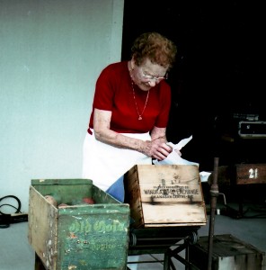 Photo en couleur d’une vieille femme debout sur une terrasse en train d’envelopper une pomme en bois avant de la déposer dans une des deux caisses de pommes placées devant elle. Elle a des cheveux roux frisés et porte un chandail rouge.