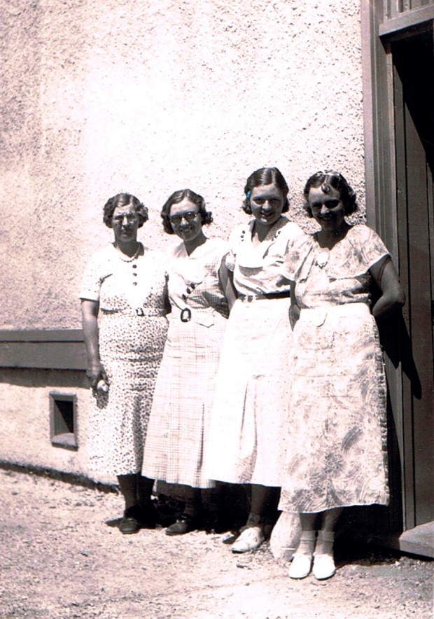 Photo en noir et blanc représentant quatre femmes, dehors, au soleil, le dos appuyé à un édifice, souriant au photographe. Elles portent toutes des robes et leurs cheveux sont coiffés à la mode des années 1930.