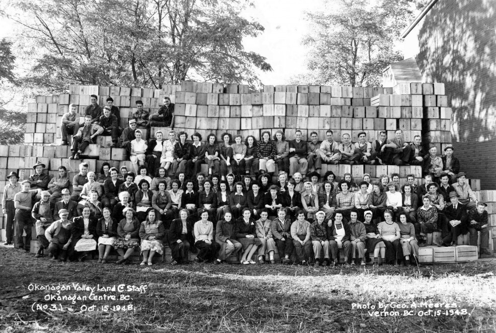 Photo officielle en noir et blanc représentant un grand groupe de femmes et d’hommes, à l’extérieur, sur des gradins, avec des caisses de pommes en arrière-plan.