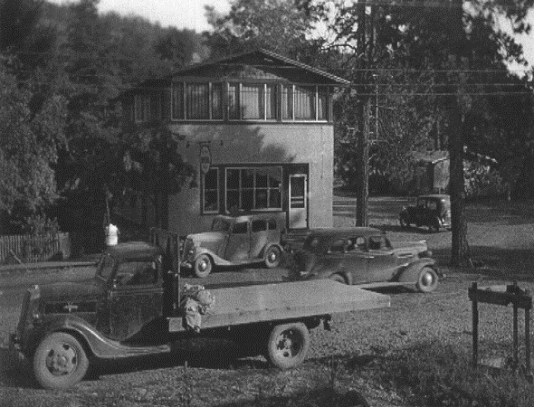 Photo en noir et blanc représentant un édifice devant lequel se trouvent un camion et trois voitures d’autrefois.