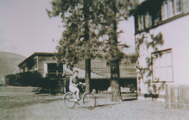 Photo en noir et blanc d’une jeune femme sur son vélo. Un édifice de bois avec une porte de chargement se dresse à l’arrière-plan.