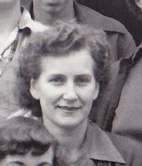 Portrait en noir et blanc d’une jeune femme souriant au photographe. Ses cheveux bouclés sont coiffés en arrière.