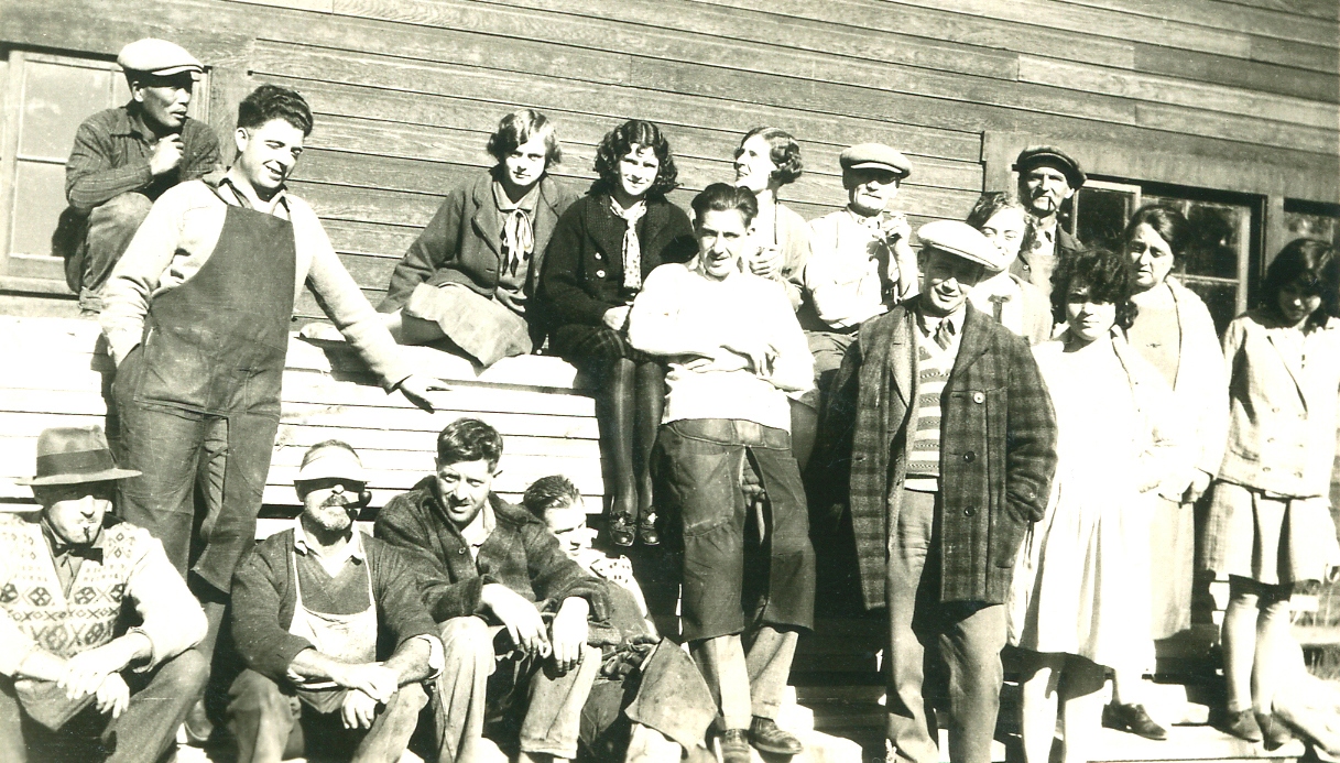 Photo spontanée en noir et blanc représentant dix hommes entre 20 et 45 ans et sept jeunes femmes portant tous des tenues décontractées, devant un édifice en bois. Certains d’entre eux sont assis sur des piles de bois, d’autres se tiennent debout ou penchés vers l’arrière. Deux hommes fument la pipe.