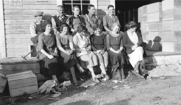 Photo en noir et blanc représentant six femmes (au premier plan) et cinq hommes (à l’arrière-plan) assis devant un vieil édifice. Ils portent des vêtements des années 1920.