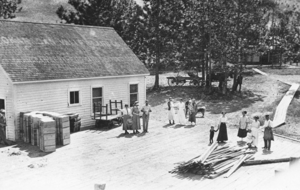 Photo en noir et blanc représentant plusieurs hommes, femmes et enfants sur une grande terrasse de bois devant un édifice blanc en bois; ils portent tous des vêtements du début du XXe siècle. Sur leur gauche, des piles de caisses de pommes et une charrette. En arrière-plan, des arbres, de l’herbe, un étroit trottoir en bois et une charrette attelée.