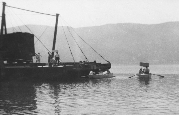 Photo en noir et blanc représentant une barge sur laquelle quatre hommes se tiennent debout. Trois personnes sont assises dans chacun des deux canots à rames qui se trouvent près de la barge. Trois personnes arborent des panneaux.